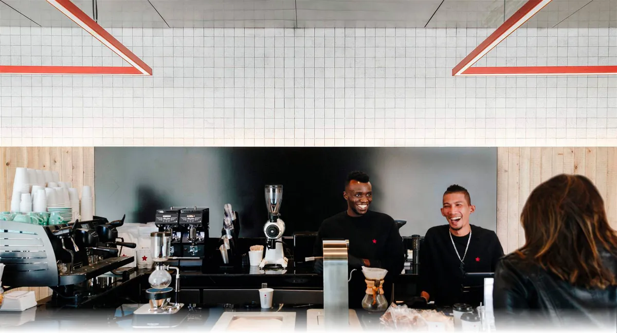 Three baristas working the espresso bar under linear red neon, guests at the counter.