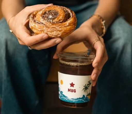 Hands holding a freshly baked cinnamon roll above an iced Americano in a branded Mug cup.